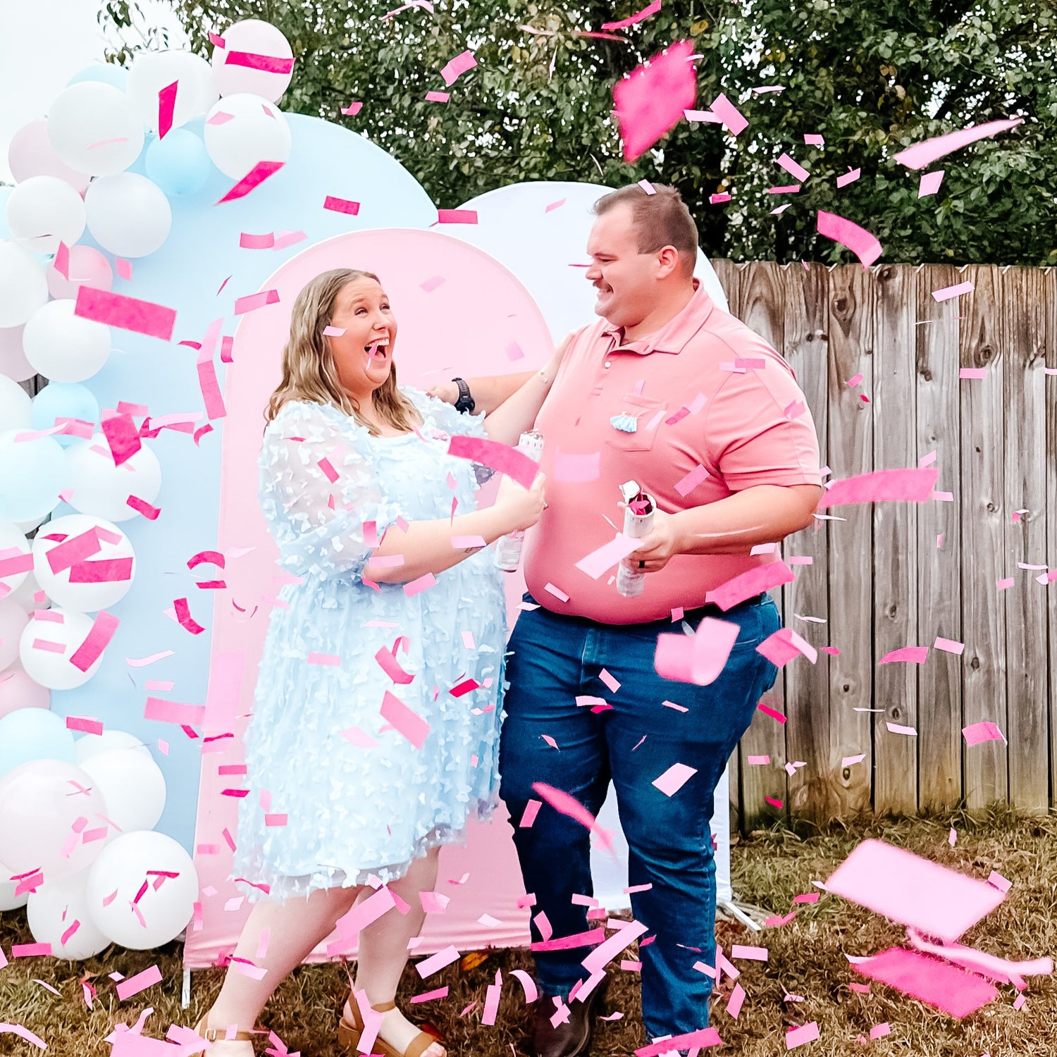 Woman in Light Blue Sheer Puff Sleeve Dress and Man in Salmon Pink Polo Shirt and Jeans Popping Pink Confetti In Front of Pink and Blue Balloon Backdrop