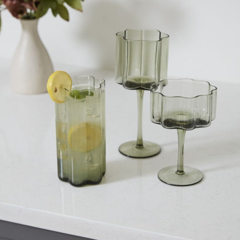 Sonnet Smoky Green Modern Floral Shape Glass Drinkware Collection, one Highball glass, one Wine glass, one Coupe glass. Highball glass filled with lemon cocktail.
