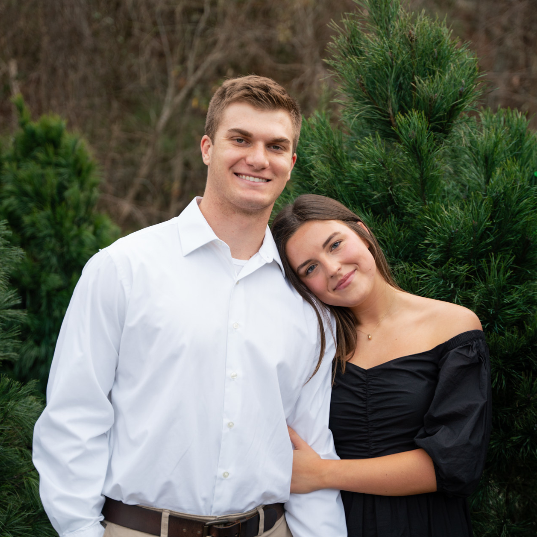 Man in White Dress Shirt and Woman in Black Off the Shoulder Dress Smiling in front of Evergreen Bushes