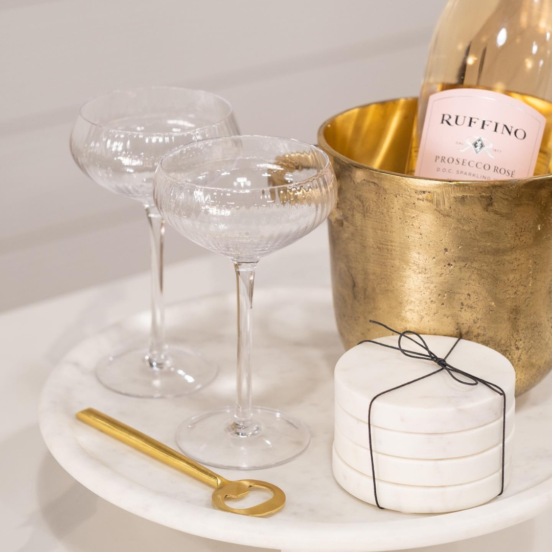 Coupe Glasses and Ice Bucket with Pink Prosecco Rosé Next to Gold Handled Bottle Opener and Stack of 4 White Marble Coasters on White Marble Pedestal